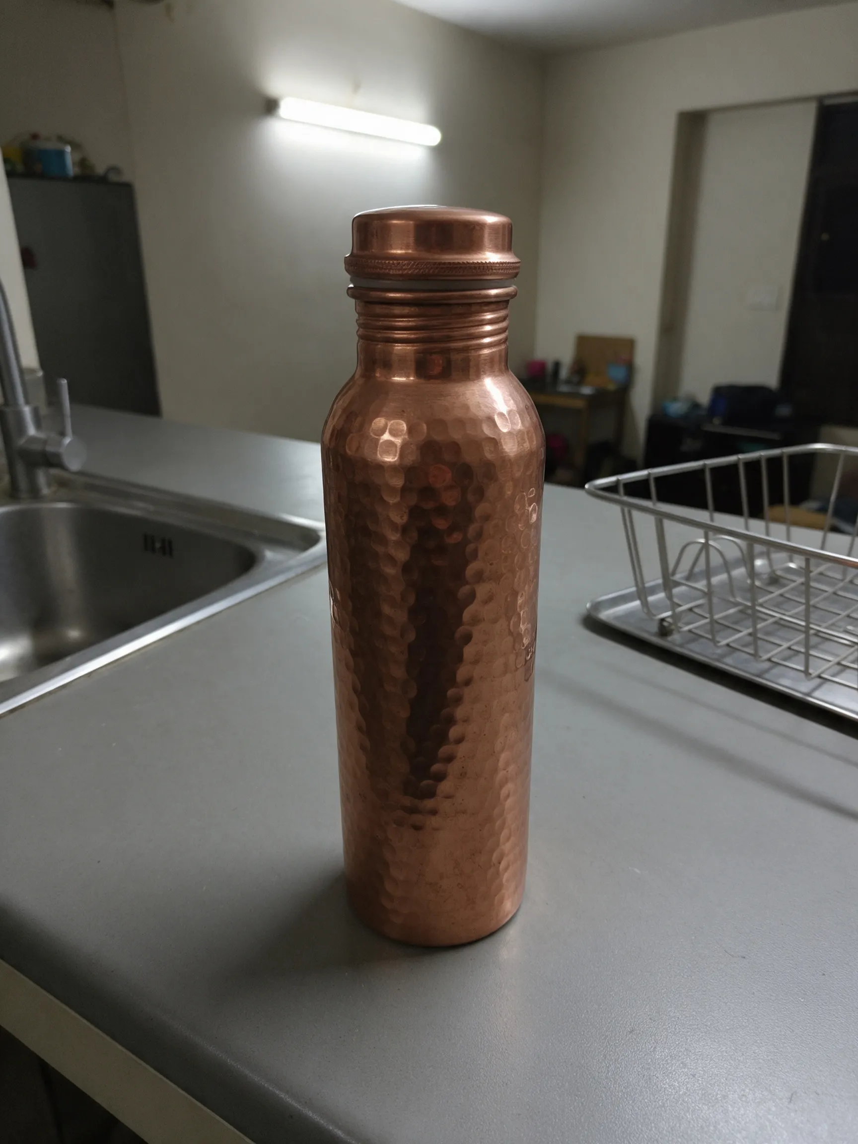 Phone photo of a hammered copper water bottle on a grey kitchen counter next to a steel sink and dish rack under tube light