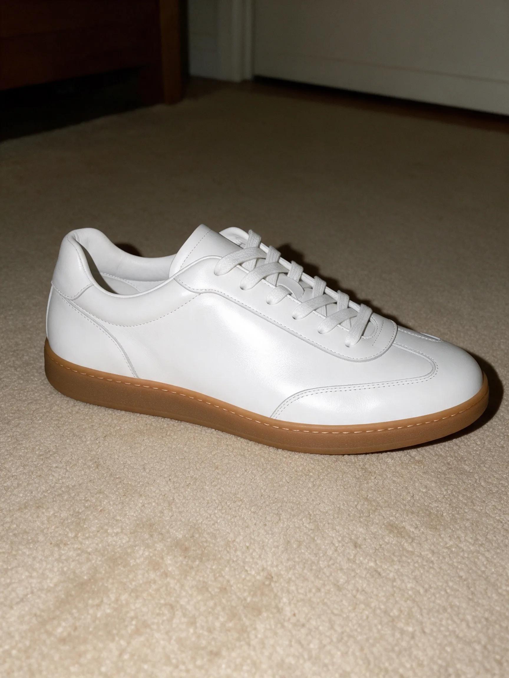 Flash phone photo of a white leather low-top sneaker with a gum sole sitting on a beige bedroom rug